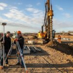 Surveyors using a total station on a construction site with a geotechnical drill rig operating in the background.
